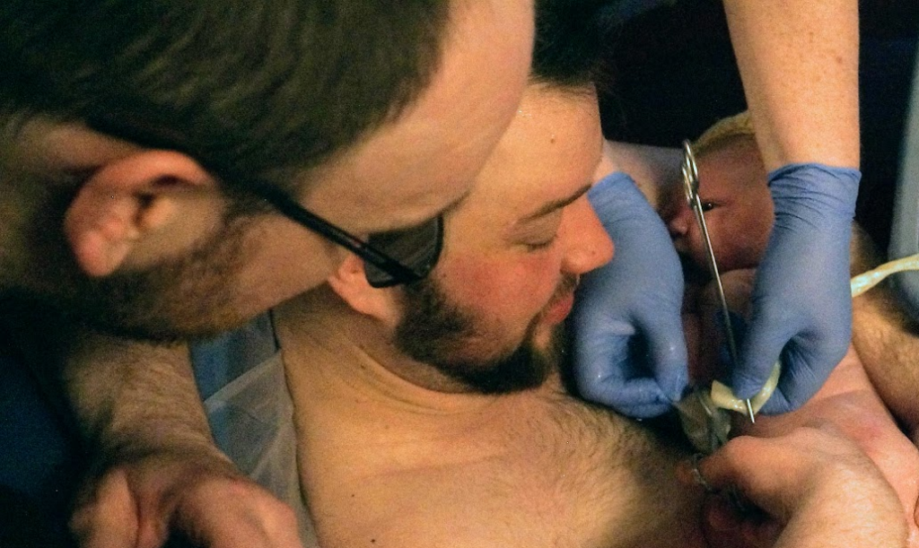 Jacob, a white trans man, is in a birthing pool holding his newborn baby in his arms. His husband Andy is leaning over to cut the umbillical cord. The midwife is holding the cord ready to be cut and she is wearing blue gloves.